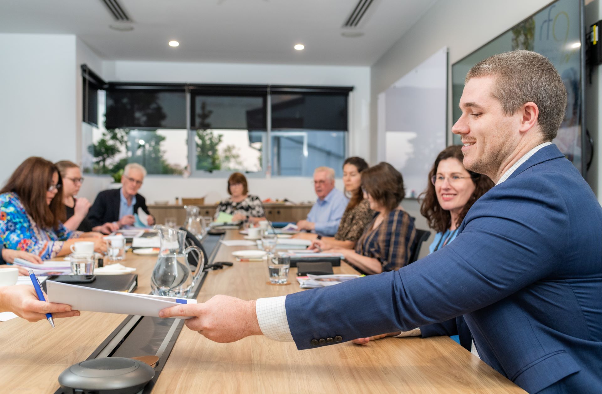 A man passes papers to a woman across a board room table